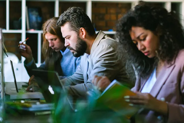 Concentrated young coworkers wearing formal clothes sitting at desk with books and working on laptops in modern coworking studio. How to schedule coworking space bookings without delays